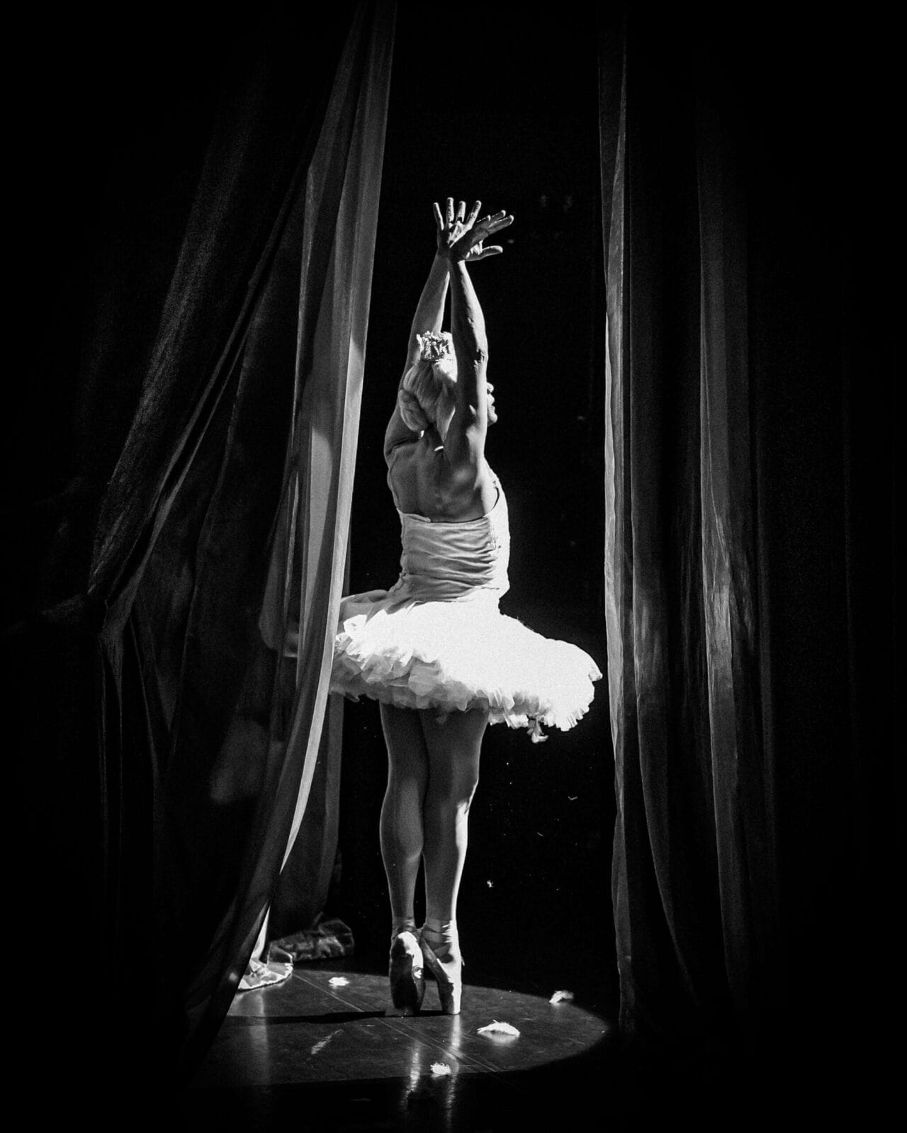 A ballerina in a tutu stands en pointe with arms raised, facing a dark stage and framed by curtains—dramatic light revealing the poise of a Monte Carlo ballet troupe dancer from behind.