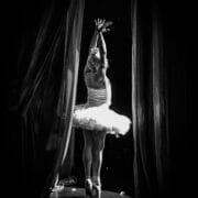 A ballerina in a tutu stands en pointe with arms raised, facing a dark stage and framed by curtains—dramatic light revealing the poise of a Monte Carlo ballet troupe dancer from behind.