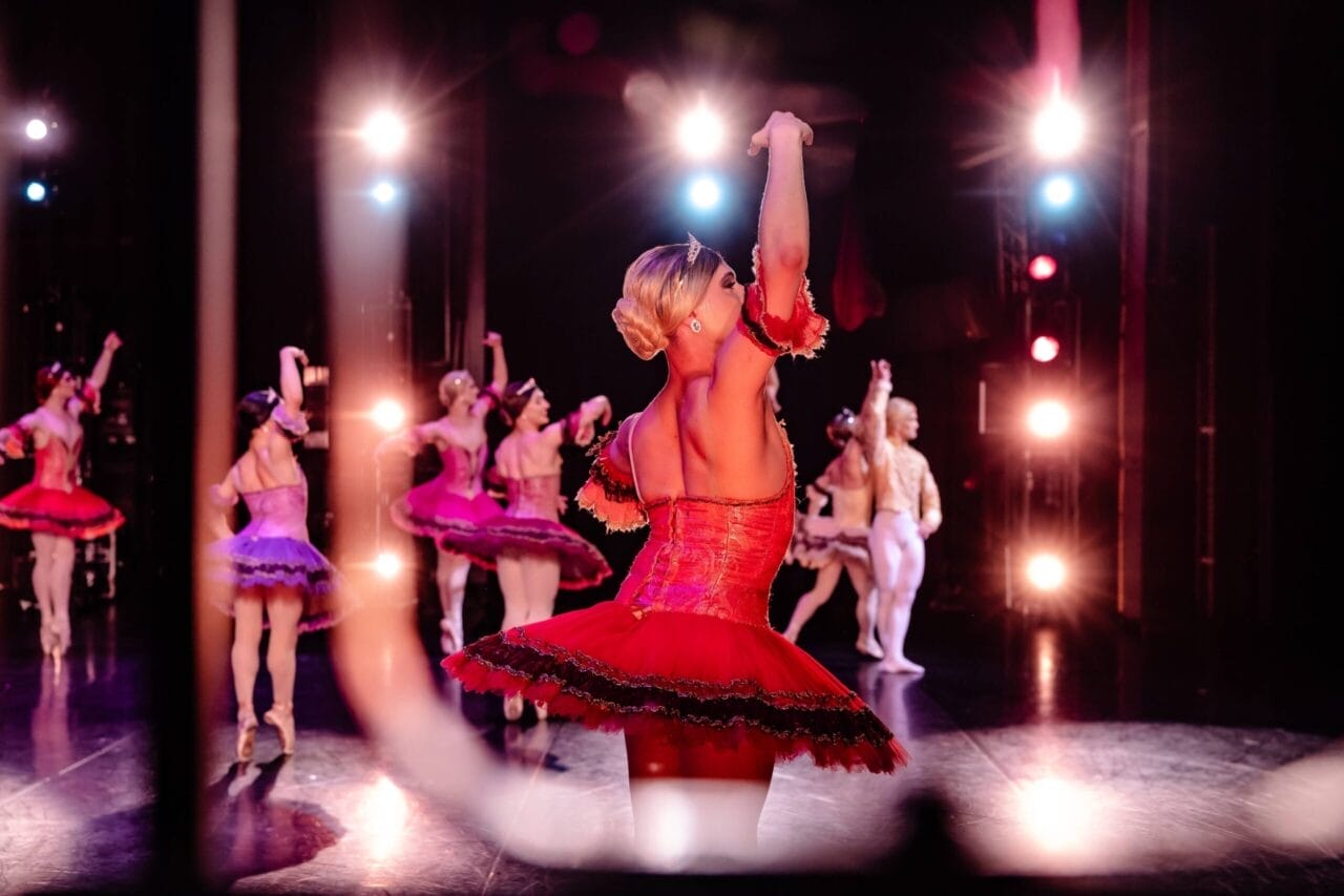 A group of ballet dancers performs on stage, with the central dancer in a bright red tutu facing away from the camera. Stage lights illuminate the scene, evoking the elegance of Monte Carlo and casting colourful reflections on the floor.
