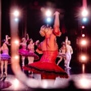 A group of ballet dancers performs on stage, with the central dancer in a bright red tutu facing away from the camera. Stage lights illuminate the scene, evoking the elegance of Monte Carlo and casting colourful reflections on the floor.