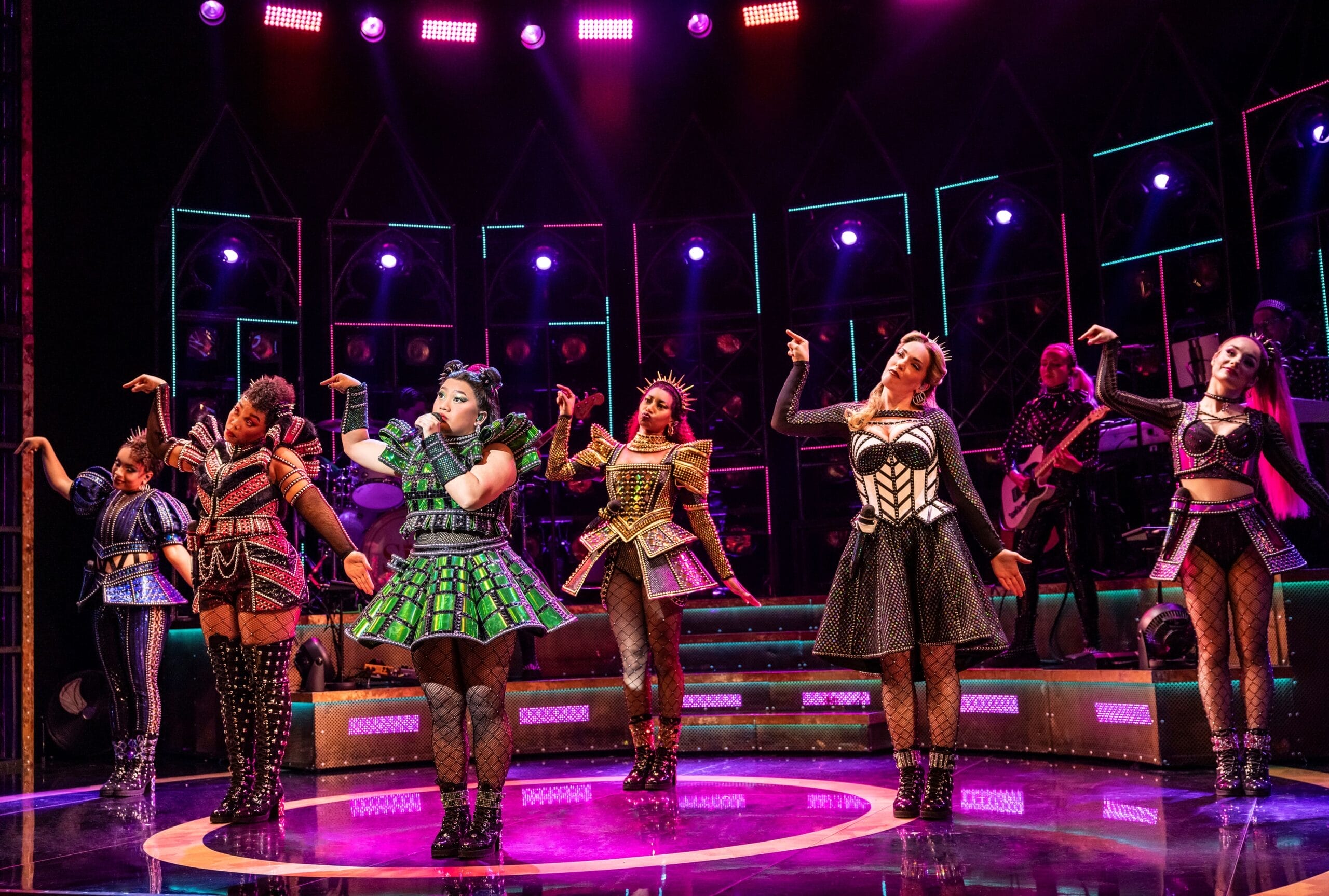 Six women in colorful, stylized costumes perform a choreographed dance on stage under vibrant lighting, with musicians playing in the background.