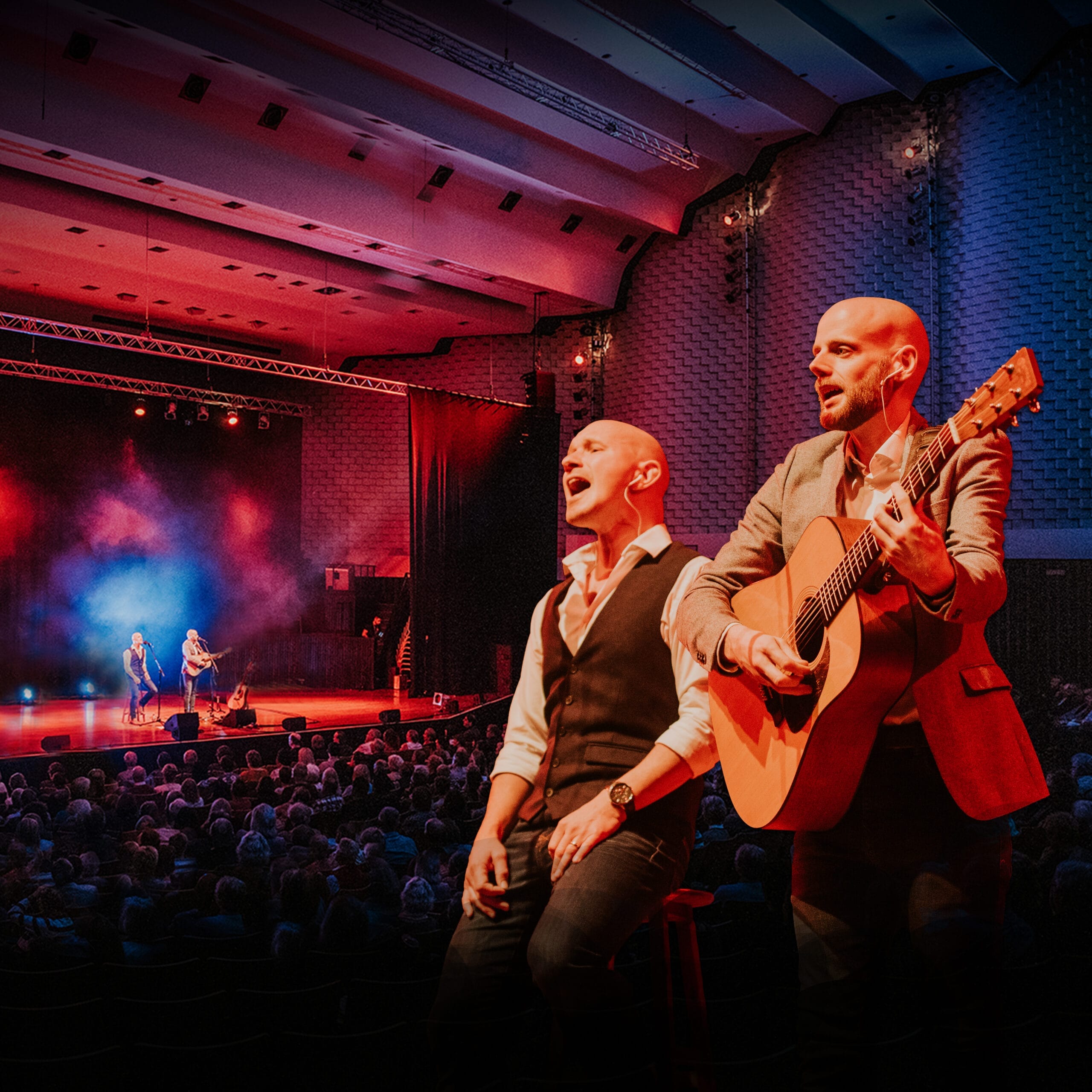 Two musicians perform on stage in a large, dimly lit concert hall with red and purple lighting—one sings seated, the other plays guitar standing—creating a Simon and Garfunkel-like atmosphere that echoes through the years.