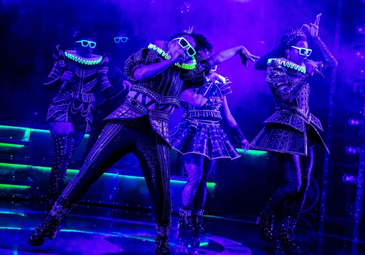 Performers in studded costumes and glowing accessories dance under purple stage lights.