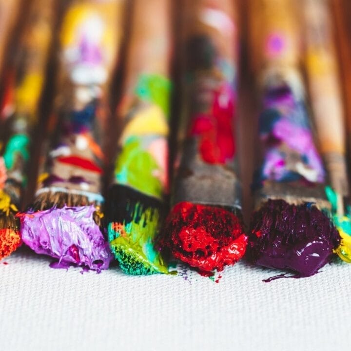 A row of paintbrushes with colorful paint on the bristles rests on a white canvas.