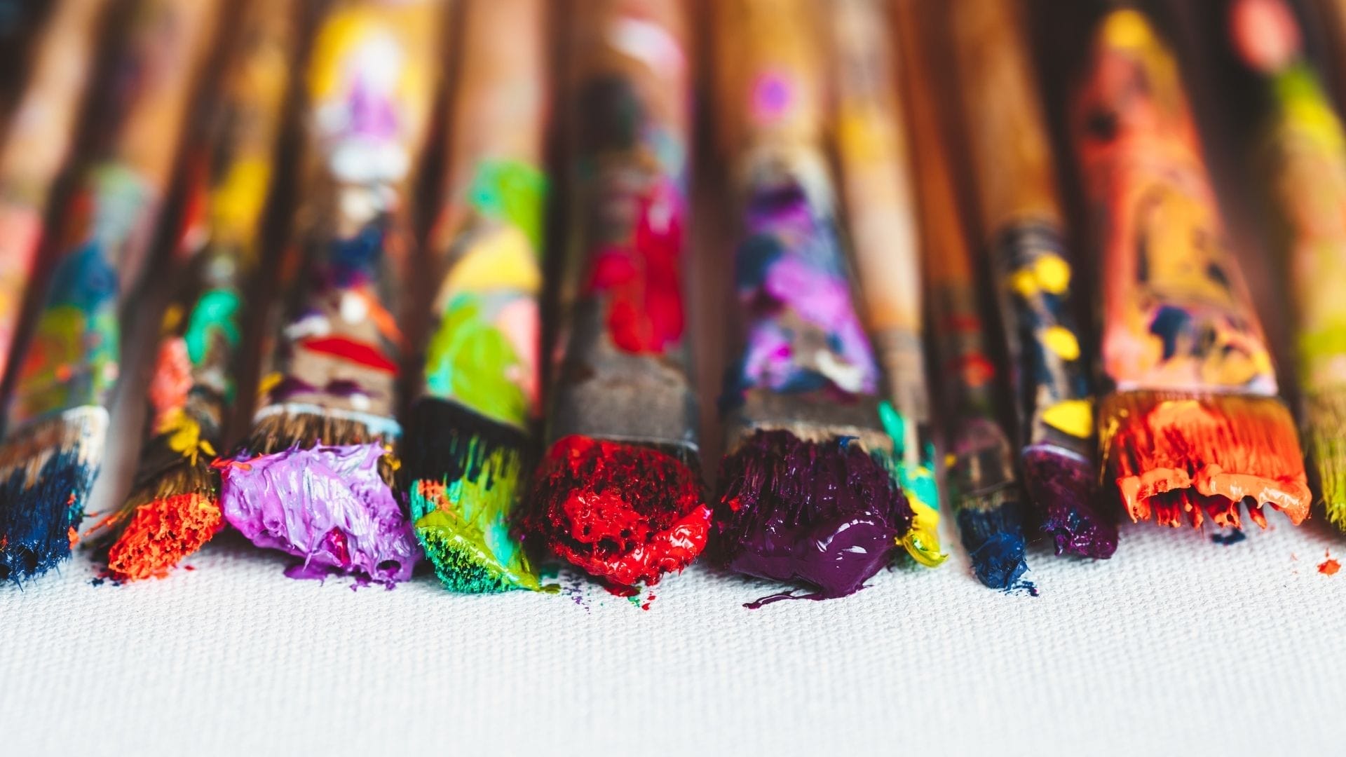 A row of paintbrushes with colorful paint on the bristles rests on a white canvas.