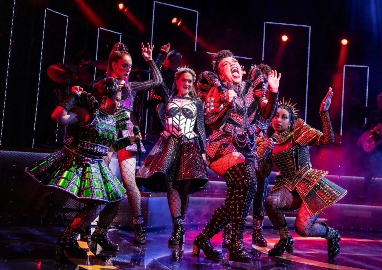 Five performers in ornate, colorful costumes sing and dance energetically on a stage with dramatic red lighting and musicians in the background.