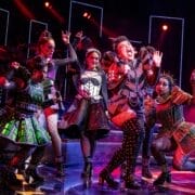 Five performers in ornate, colorful costumes sing and dance energetically on a stage with dramatic red lighting and musicians in the background.