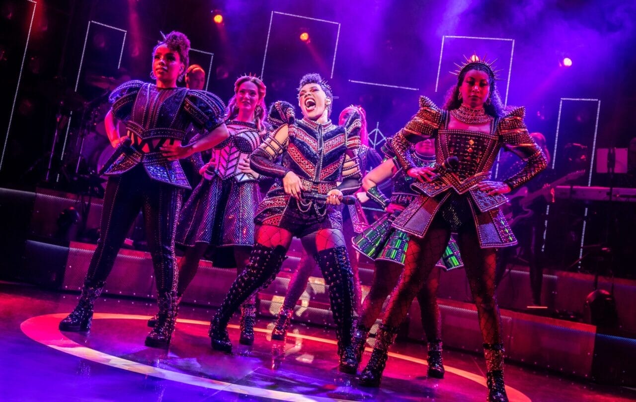 Performers in colorful, illuminated costumes sing and dance energetically on stage under dramatic lighting during a musical theater production.