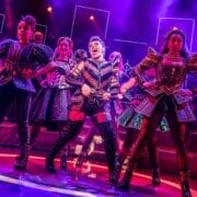 Performers in colorful, illuminated costumes sing and dance energetically on stage under dramatic lighting during a musical theater production.