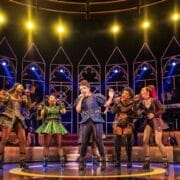 Six performers in colorful, ornate costumes sing and dance on a brightly lit stage with a cathedral-like backdrop and band visible in the background.