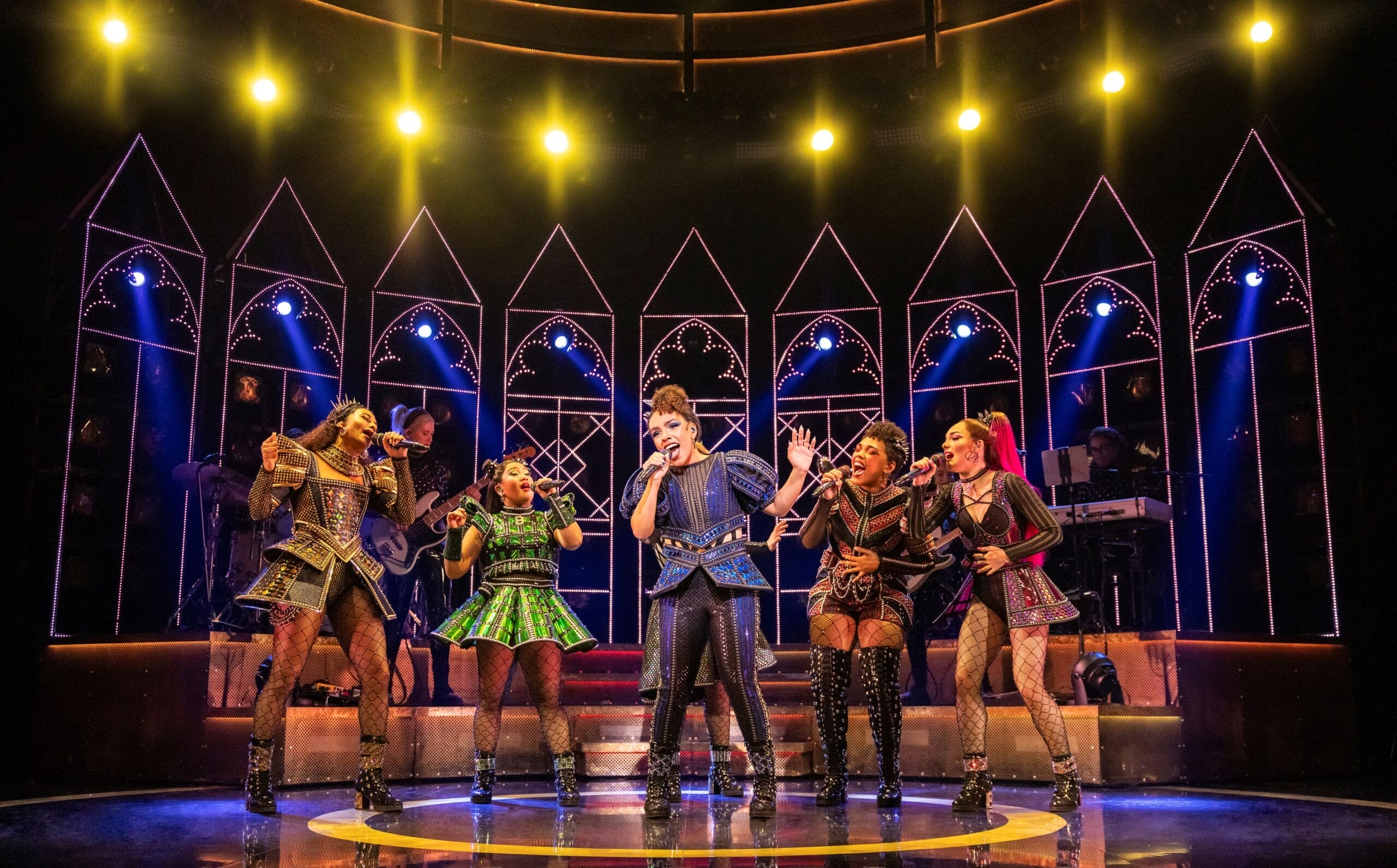 Six performers in colorful, ornate costumes sing and dance on a brightly lit stage with a cathedral-like backdrop and band visible in the background.