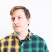 A person with short light brown hair wears a half yellow, half green plaid shirt, standing against a plain white background, evoking the vibrant spirit of musical theatre.