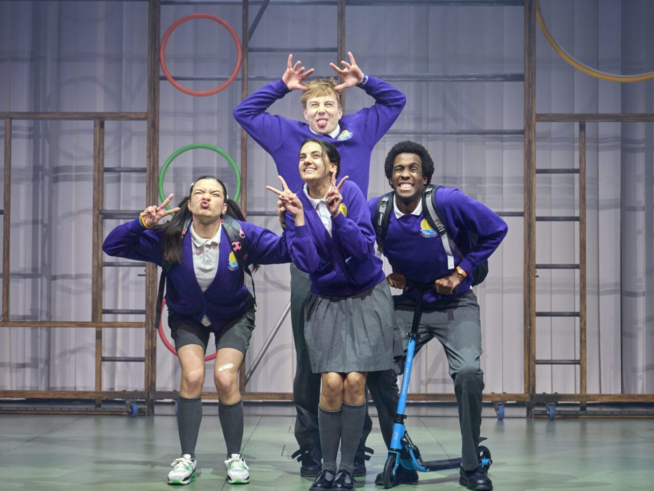 Four young people in school uniforms pose playfully on stage, reminiscent of characters from a children's book. Three stand, pulling funny faces and making hand gestures, while one kneels on a scooter beneath colourful hoops.