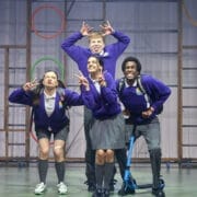 Four young people in school uniforms pose playfully on stage, reminiscent of characters from a children's book. Three stand, pulling funny faces and making hand gestures, while one kneels on a scooter beneath colourful hoops.