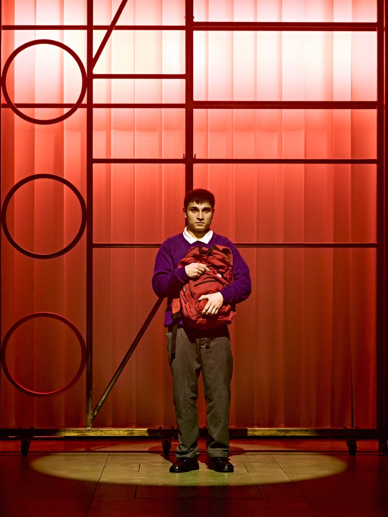 A young man stands centre stage against a red-lit background, wearing a purple jumper, white shirt, and grey trousers—evoking the spirit of The Boy at the Back of the Class by Onjali Q. Raúf—as he holds a red item of clothing under a spotlight.