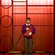 A young man stands centre stage against a red-lit background, wearing a purple jumper, white shirt, and grey trousers—evoking the spirit of The Boy at the Back of the Class by Onjali Q. Raúf—as he holds a red item of clothing under a spotlight.