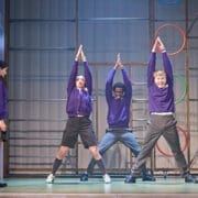Four students in school uniforms perform a synchronised movement on stage, as if acting out a scene from the bestselling novel The Boy at the Back of the Class. Coloured rings are mounted on a metal structure in the background.