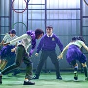 Four young actors in school uniforms perform energetically onstage, bringing to life Onjali Q Raúf’s "The Boy at the Back of the Class." The backdrop features metal bars and hanging coloured rings, echoing scenes from the beloved children’s book.