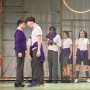 Six young people in school uniforms stand on a stage, evoking the spirit of Onjali Q. Raúf’s children’s book, The Boy at the Back of the Class. Two boys face each other closely as four others watch, with colourful rings and a wooden frame set behind them.