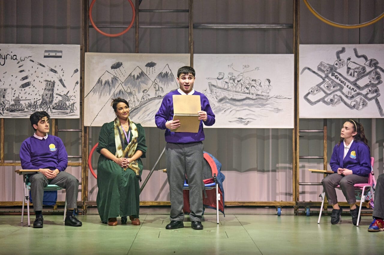 Four people sit on stage, three in purple school uniforms and one in a green dress and scarf, bringing Onjali Q. Raúf’s children’s book The Boy at the Back of the Class to life. Behind them are black-and-white sketches of maps, mountains, and a boat of people.