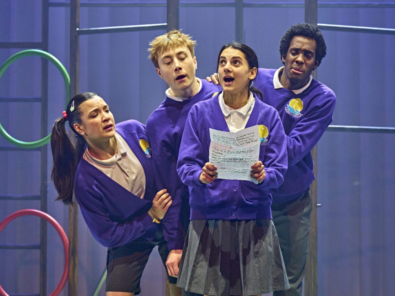 Four young actors in purple school uniforms perform on stage, bringing Onjali Q. Raúf’s children’s book "The Boy at the Back of the Class" to life. One sings from a handwritten paper as others gather close, with gym equipment in the background.