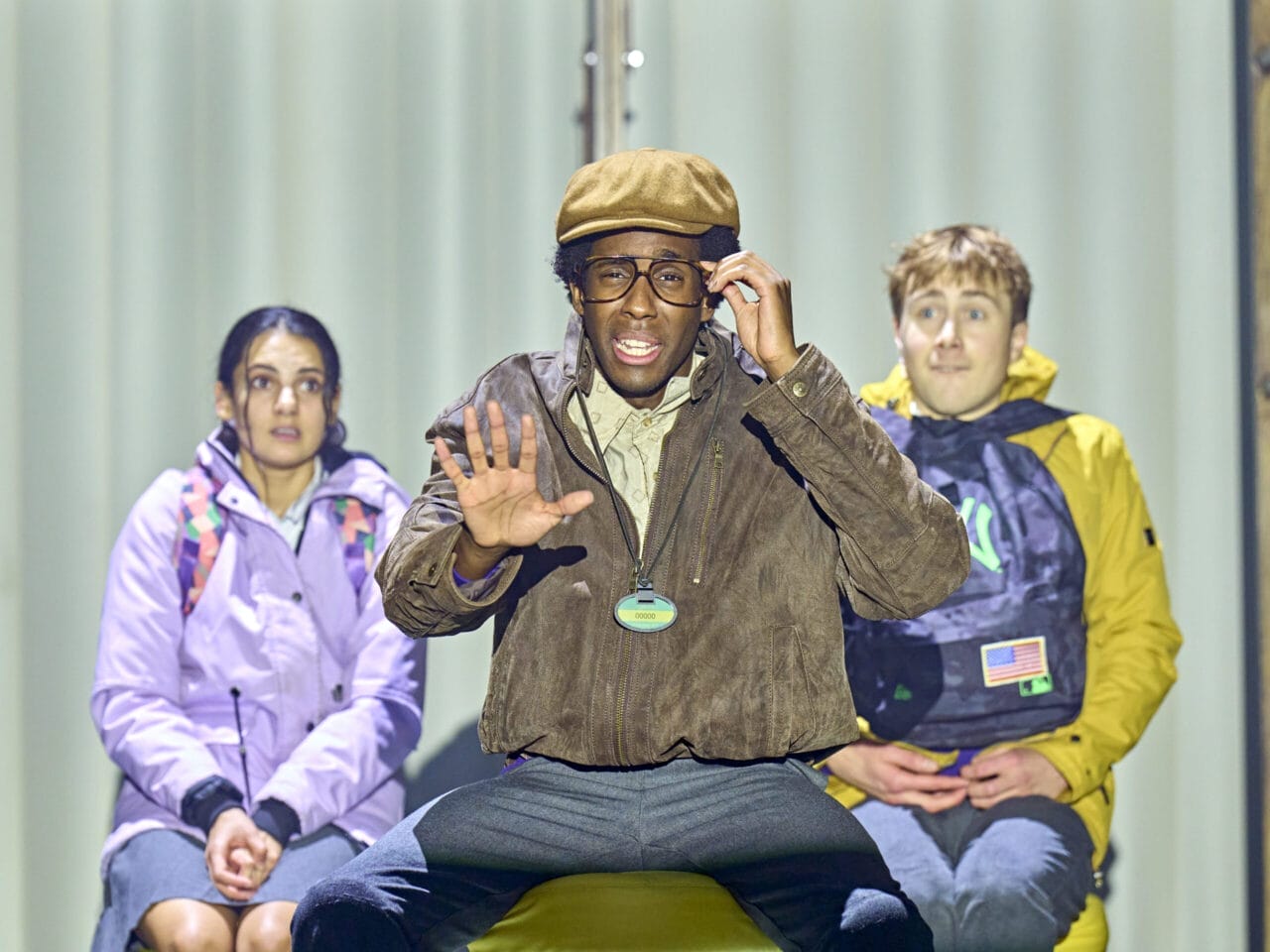 Three people sit side by side; the one in the middle—reminiscent of The Boy at the Back of the Class—wears a brown jacket, glasses, and a cap, raising one hand. The others wear jackets and rucksacks, appearing slightly anxious in a well-lit setting.