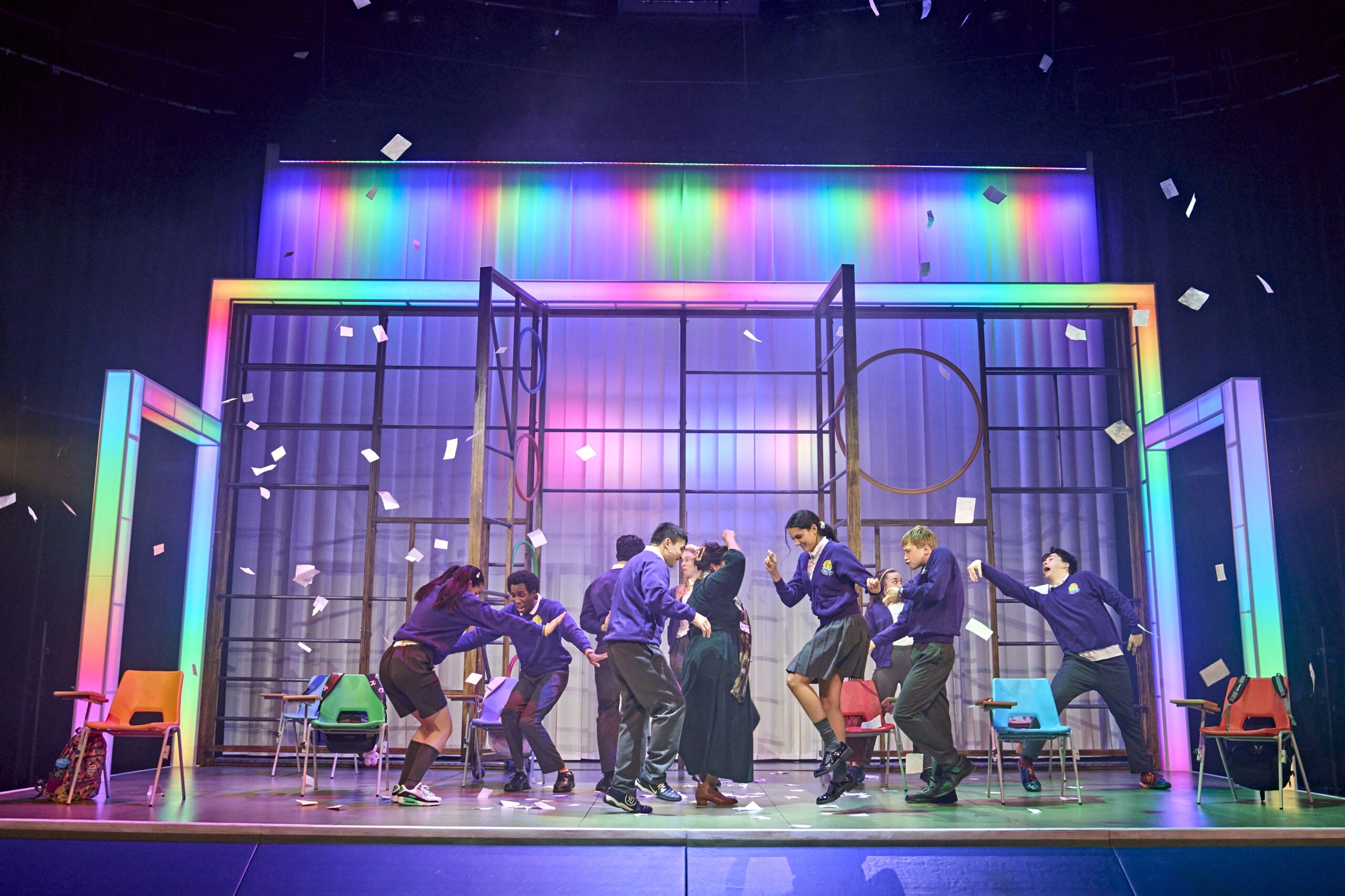 A group of people in purple jumpers and dark skirts or trousers stand on stage with colourful lights and flying papers, bringing to life scenes from Onjali Q. Raúf’s children’s book, The Boy at the Back of the Class. Some are mid-motion; chairs are scattered behind them.