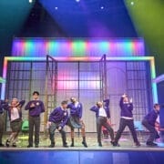 A group of performers in school uniforms pose energetically on stage before a colourful, illuminated set inspired by the children's book The Boy at the Back of the Class, while a woman in dark clothing stands at the far right.