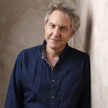A middle-aged man with gray hair wearing a navy button-up shirt leans against a beige textured wall, looking at the camera with a slight smile reminiscent of Glynis Barber’s calm confidence.
