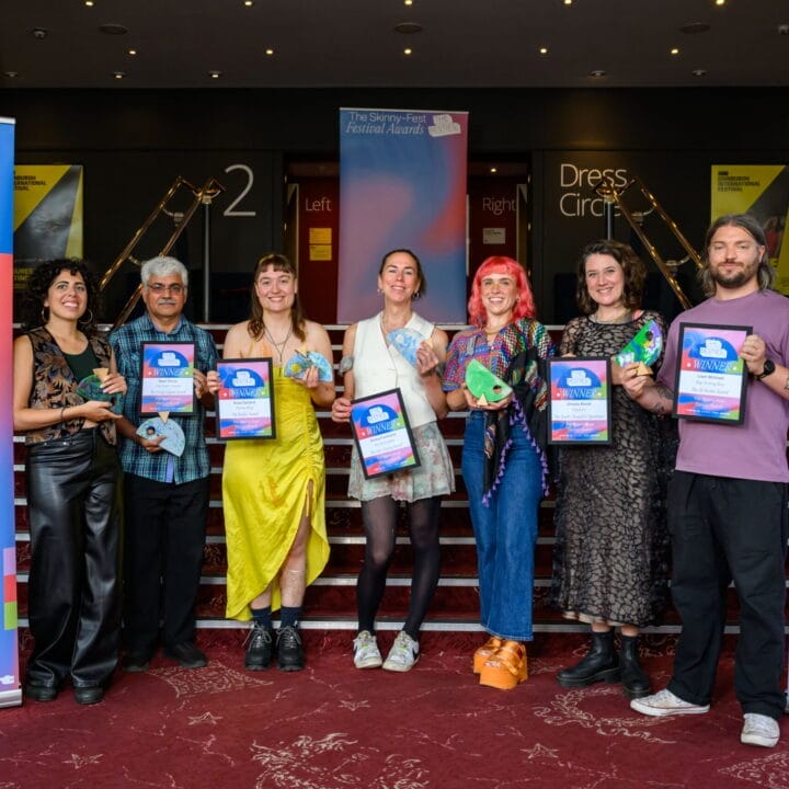 Six artists stand in a row on a red carpet, holding colorful plaques and certificates as final week winners at "The Besties Awards," with banners for the awards displayed proudly on either side.