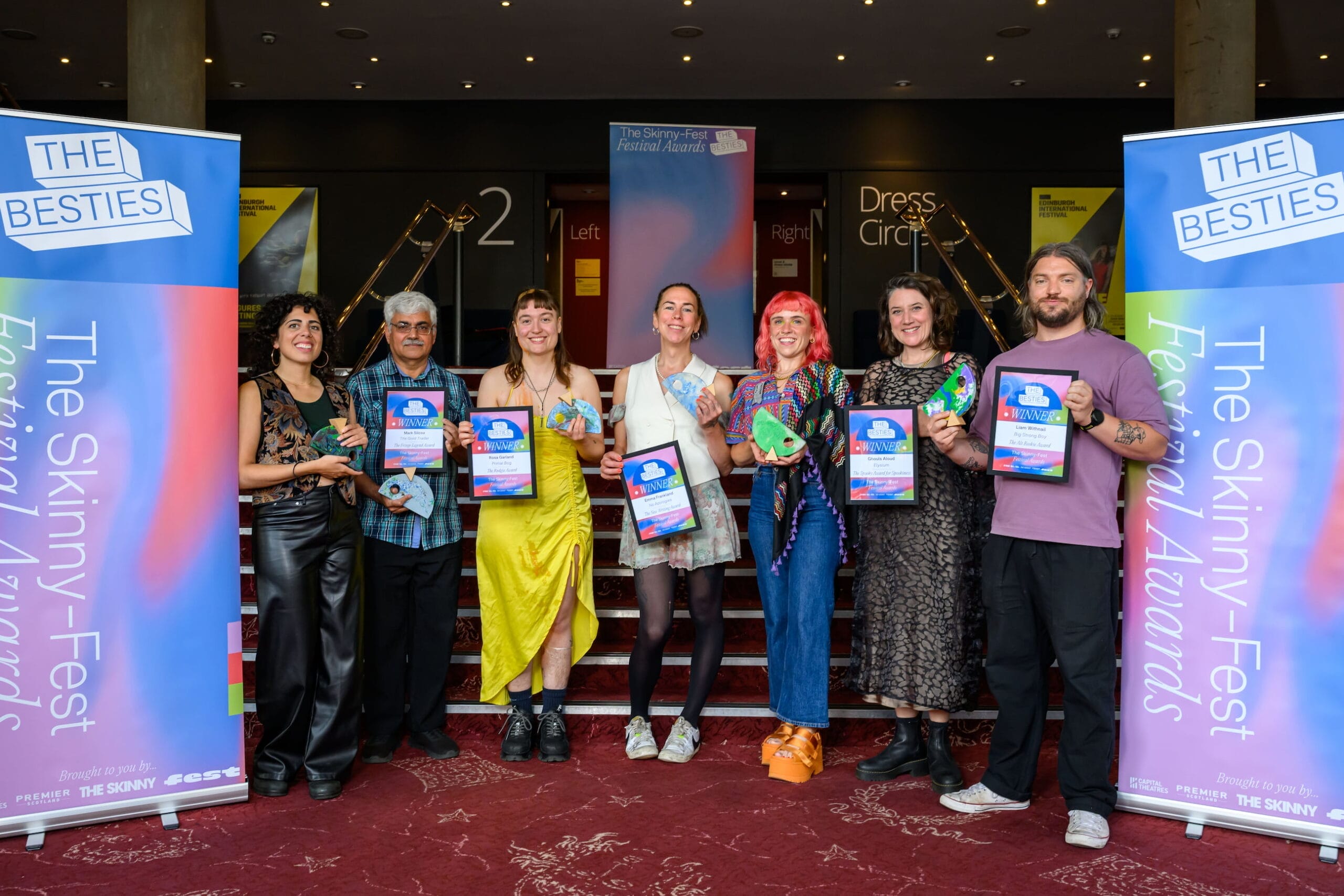 Six artists stand in a row on a red carpet, holding colorful plaques and certificates as final week winners at "The Besties Awards," with banners for the awards displayed proudly on either side.