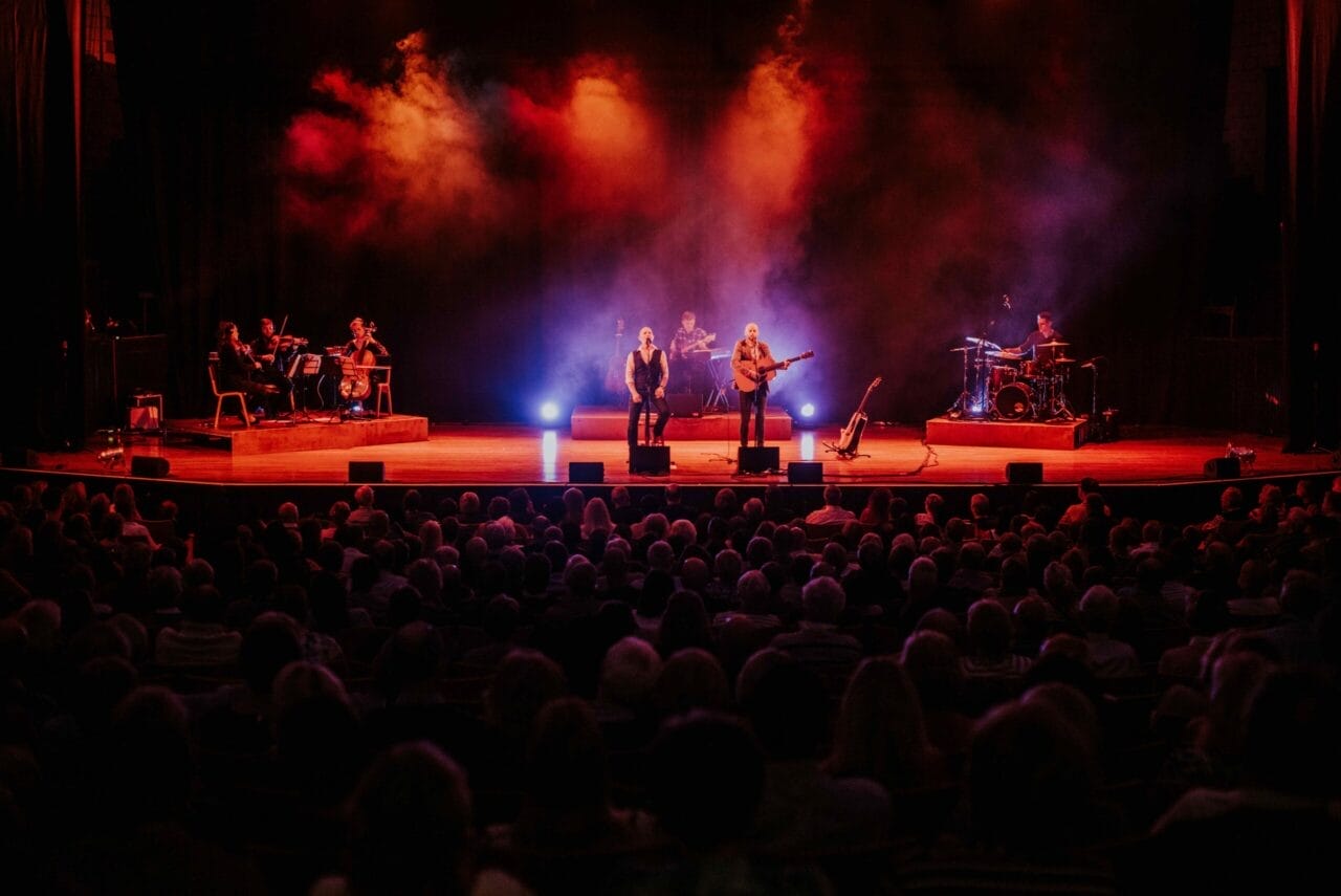 A band performs on stage with colorful lights and smoke effects, capturing the spirit of Simon and Garfunkel as they play to a seated audience in a dark theater.