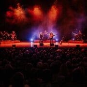 A band performs on stage with colorful lights and smoke effects, capturing the spirit of Simon and Garfunkel as they play to a seated audience in a dark theater.