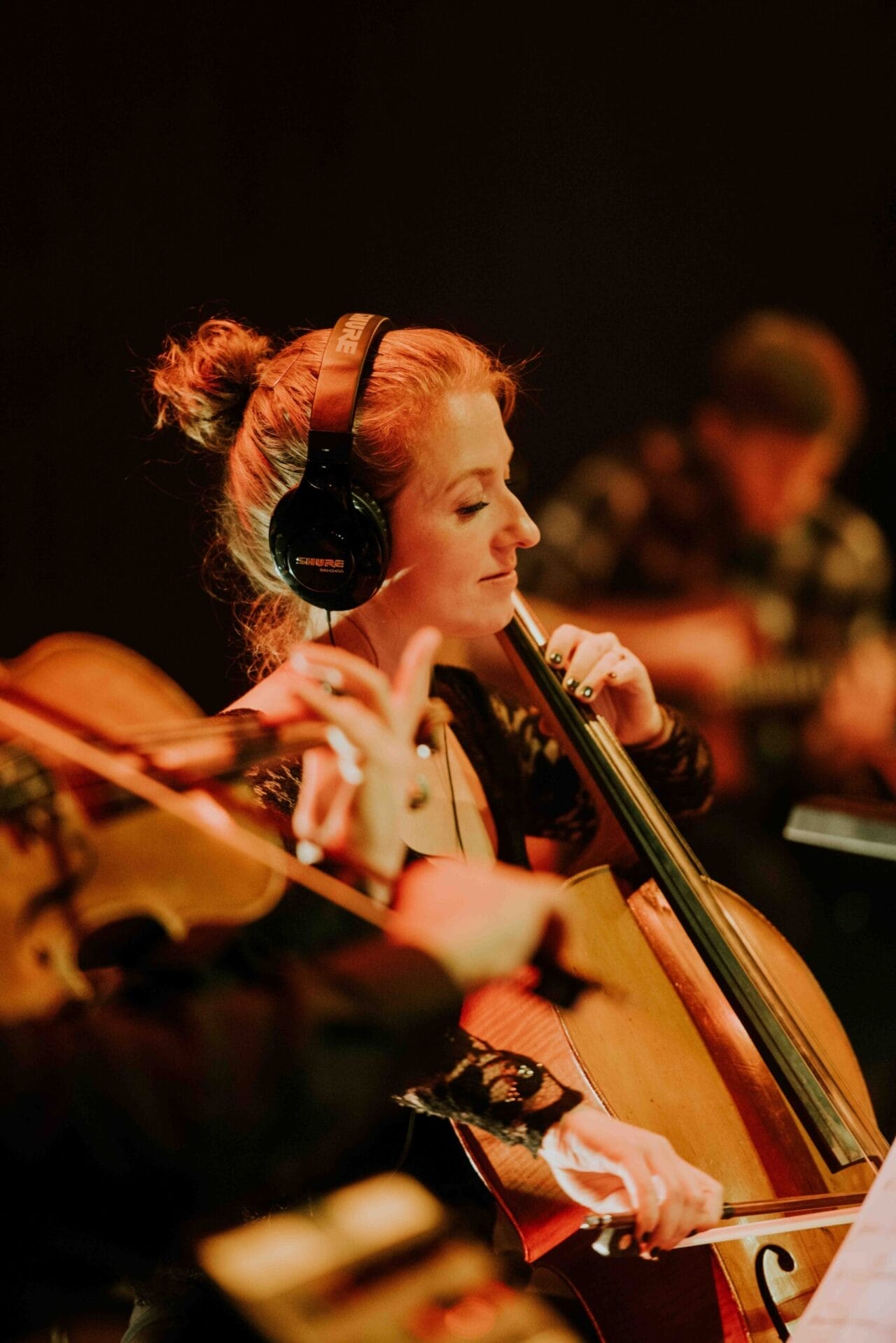 A woman wearing headphones plays the cello in an ensemble with other musicians in a dimly lit setting, evoking the timeless harmonies of Simon and Garfunkel.
