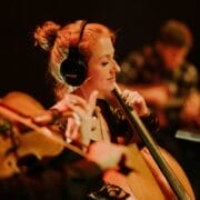A woman wearing headphones plays the cello in an ensemble with other musicians in a dimly lit setting, evoking the timeless harmonies of Simon and Garfunkel.