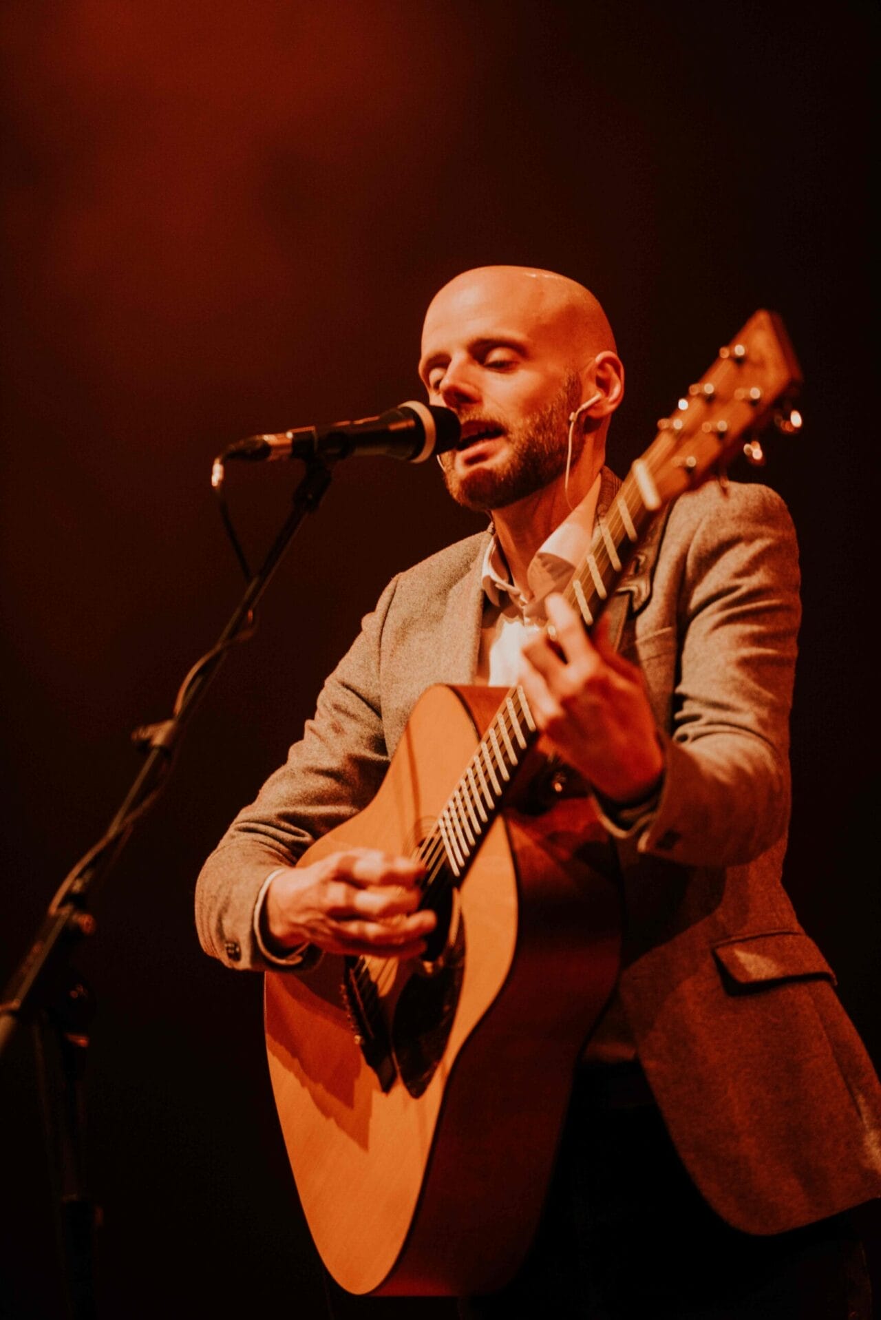A man in a gray blazer plays an acoustic guitar and sings into a microphone on stage under warm, orange lighting, delivering a heartfelt Simon and Garfunkel tribute.