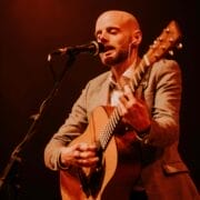 A man in a gray blazer plays an acoustic guitar and sings into a microphone on stage under warm, orange lighting, delivering a heartfelt Simon and Garfunkel tribute.