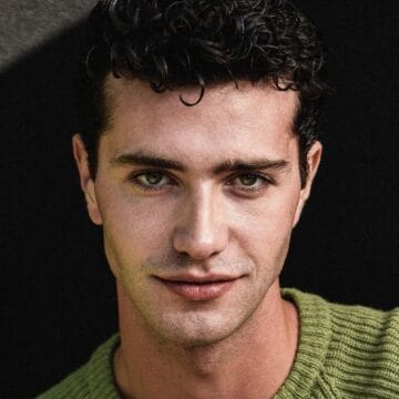 A young man with short, curly dark hair wears a green knit sweater and looks directly at the camera, set against a dark background—reminiscent of Bruce Herbelin-Earle's signature style.