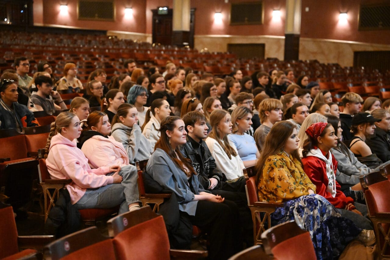 A group of young people sat in an auditorium watching and listening intently.