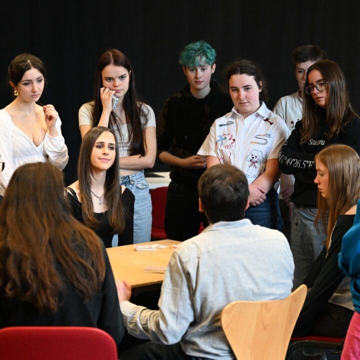 A group of people gather around a table, some seated and some standing, engaged in conversation as if rehearsing for a theatre production, in a room with a black backdrop.