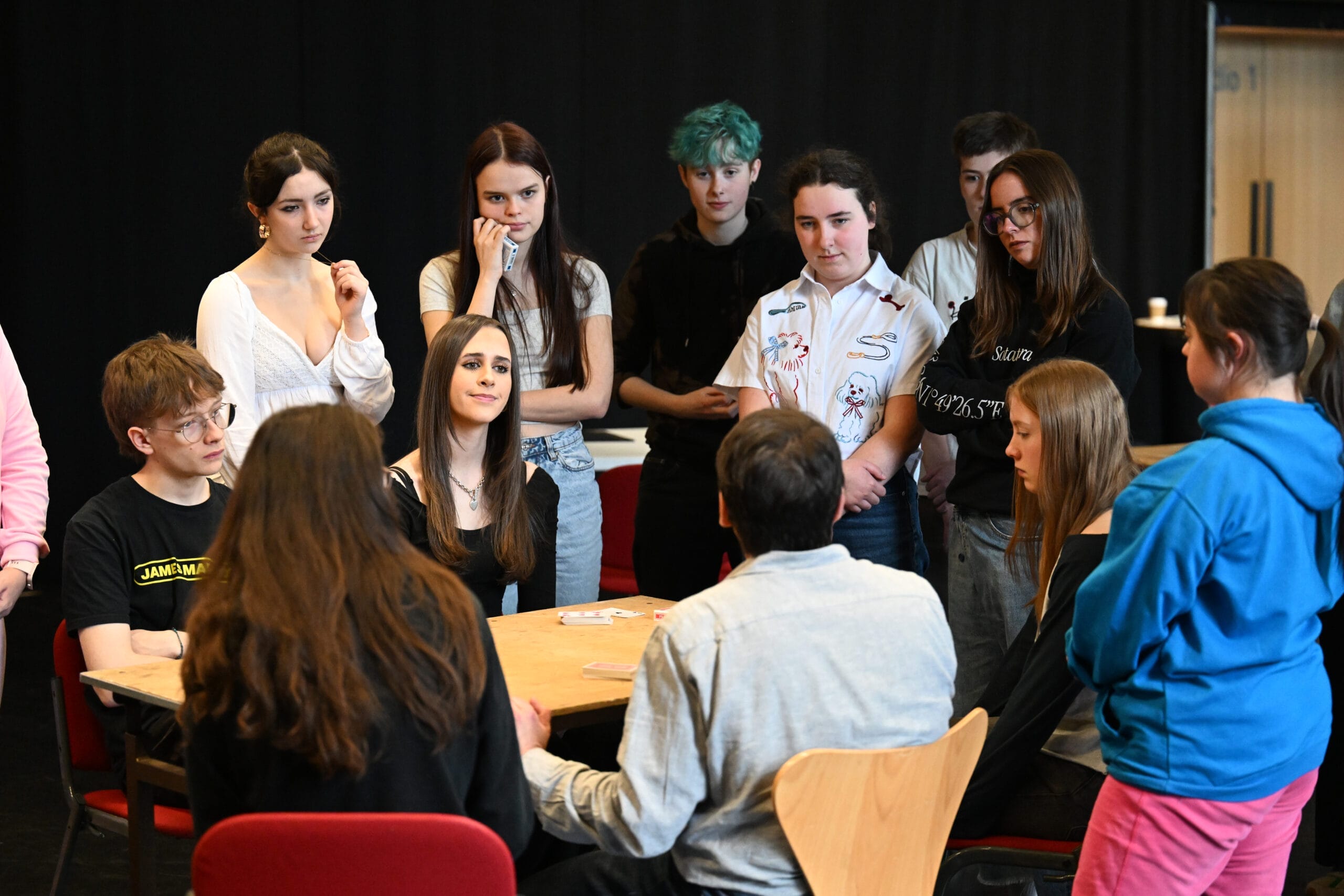 A group of people gather around a table, some seated and some standing, engaged in conversation as if rehearsing for a theatre production, in a room with a black backdrop.