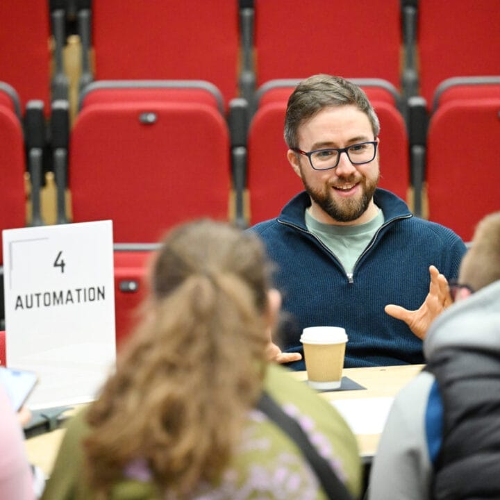A man sits at a table with three people, discussing automation during Coffee With The Creatives. He gestures with his hands and holds a coffee cup; red auditorium seats are visible in the background.