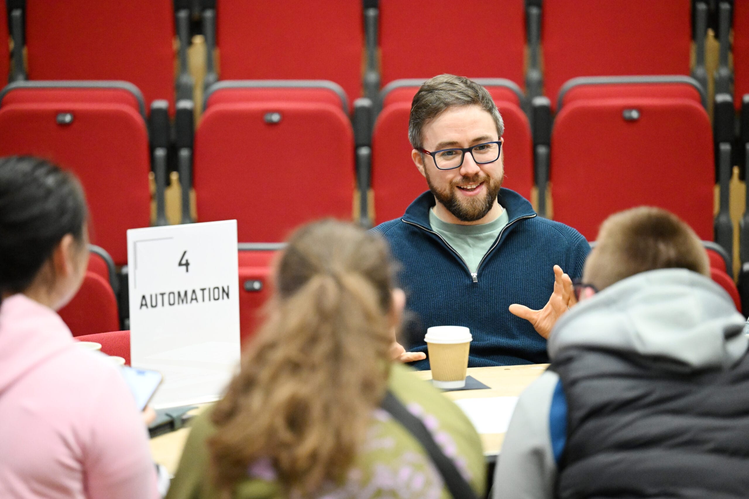 A man sits at a table with three people, discussing automation during Coffee With The Creatives. He gestures with his hands and holds a coffee cup; red auditorium seats are visible in the background.