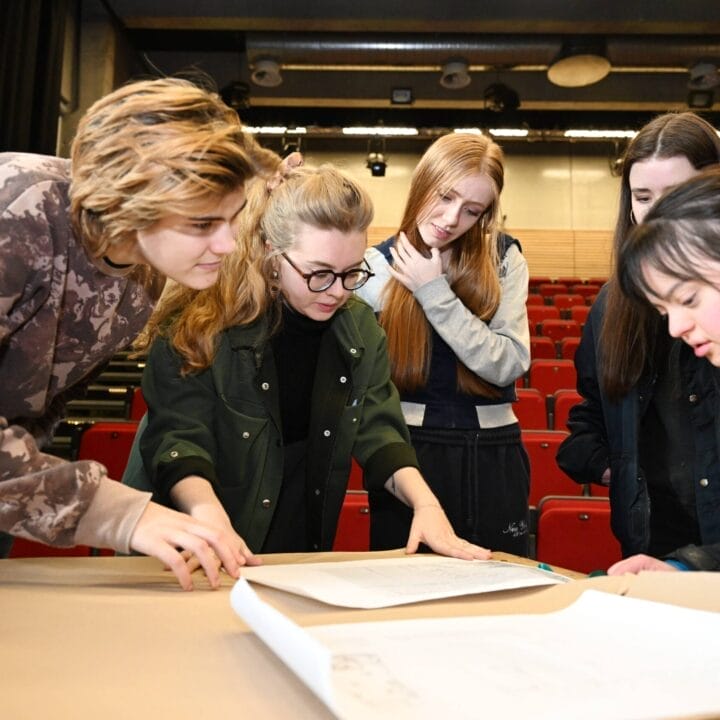 Five Student Ambassadors stand around a table in an auditorium, looking at and discussing large sheets of paper spread out before them.
