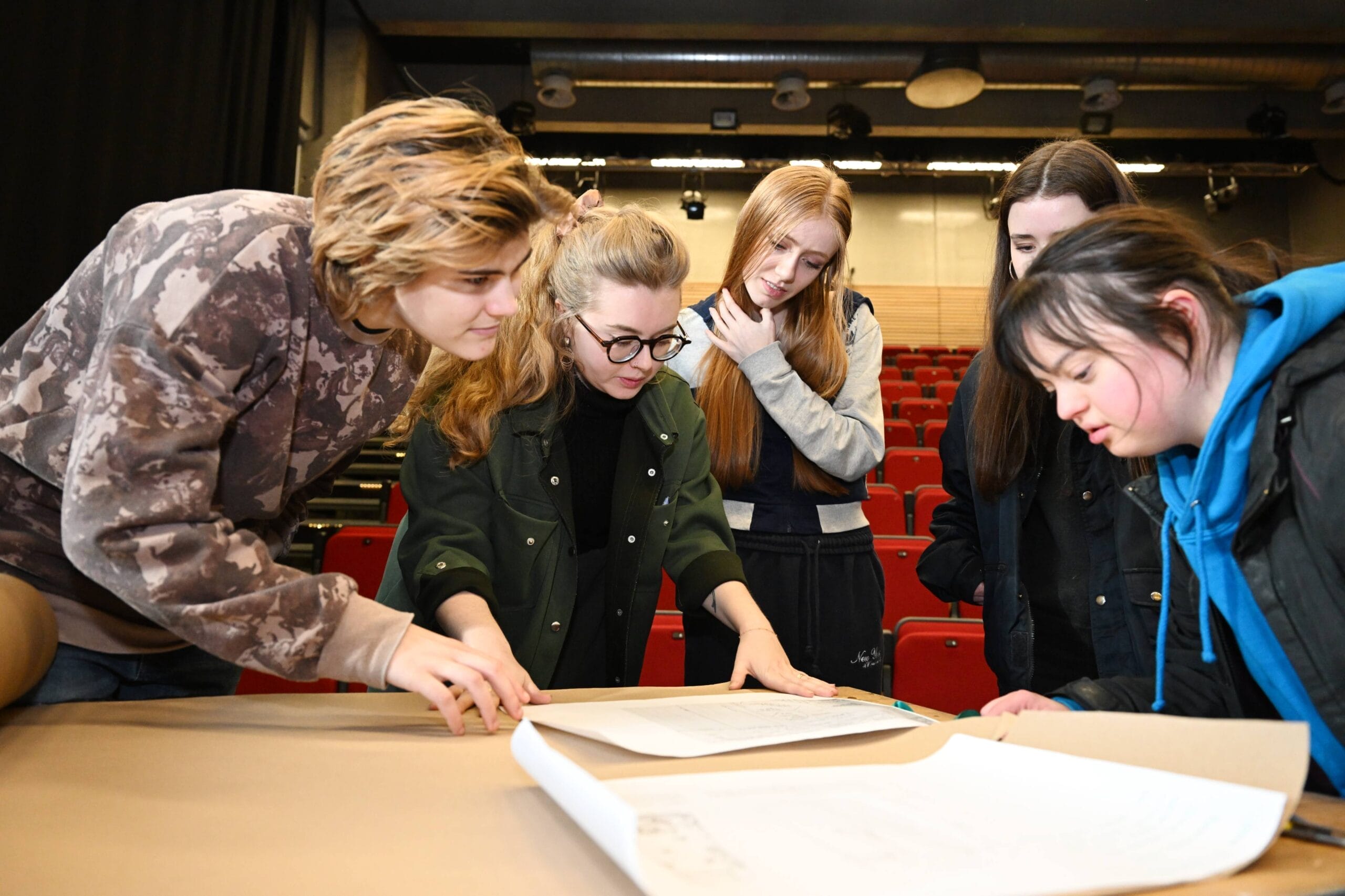 Five Student Ambassadors stand around a table in an auditorium, looking at and discussing large sheets of paper spread out before them.