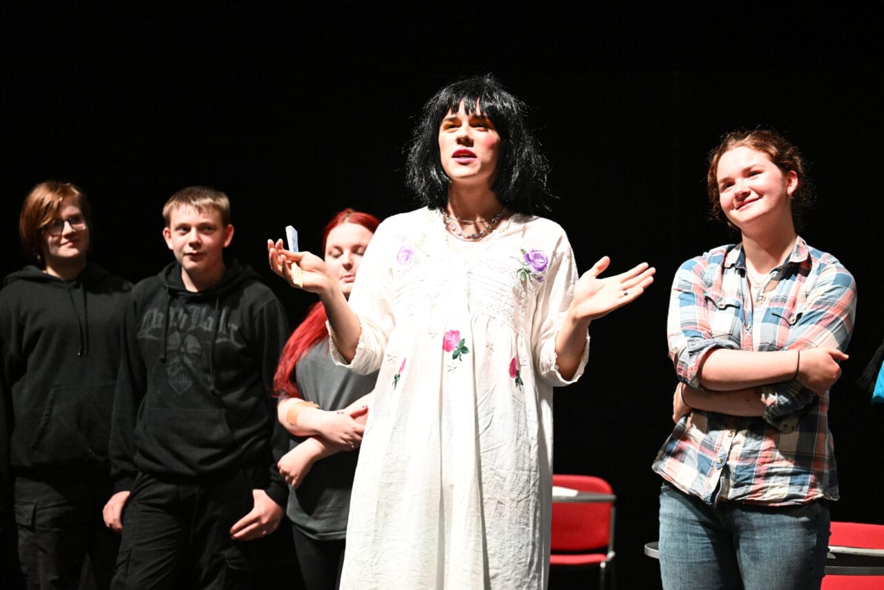 A group of young people stand on stage; one person in a white dress with a black wig gestures while four others watch and smile.