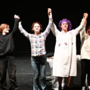 Four young people in various costumes stand, holding hands above their head readying themselves to bow.