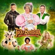 Six cast members in costume for "Jack and the Beanstalk" pose against a green background with castle elements, showcasing characters including Jack, the Dame, the Cow, and a fairy.