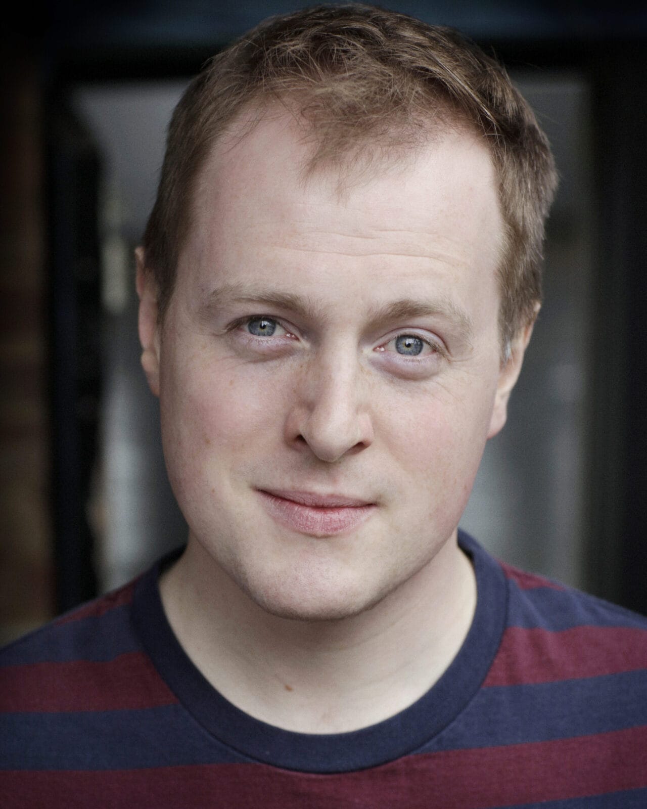 A man with short light brown hair and blue eyes is wearing a maroon and navy striped shirt, looking directly at the camera with a neutral expression.
