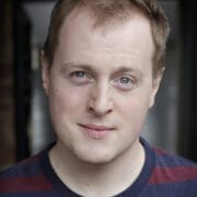 A man with short light brown hair and blue eyes is wearing a maroon and navy striped shirt, looking directly at the camera with a neutral expression.